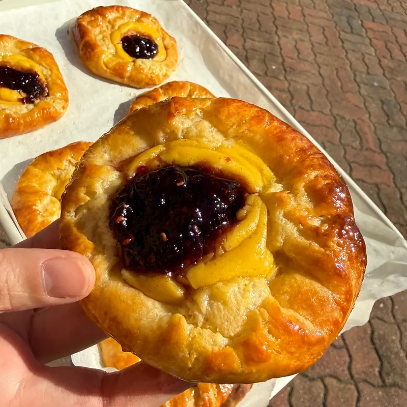 A golden Custard & Jam Danish held over a tray of more danishes